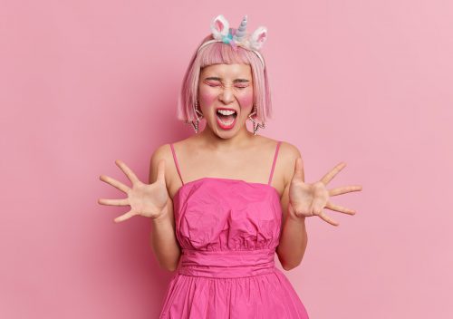 Emotional pink haired Asian woman with makeup and rhinestones on face raises palms screams angrily b