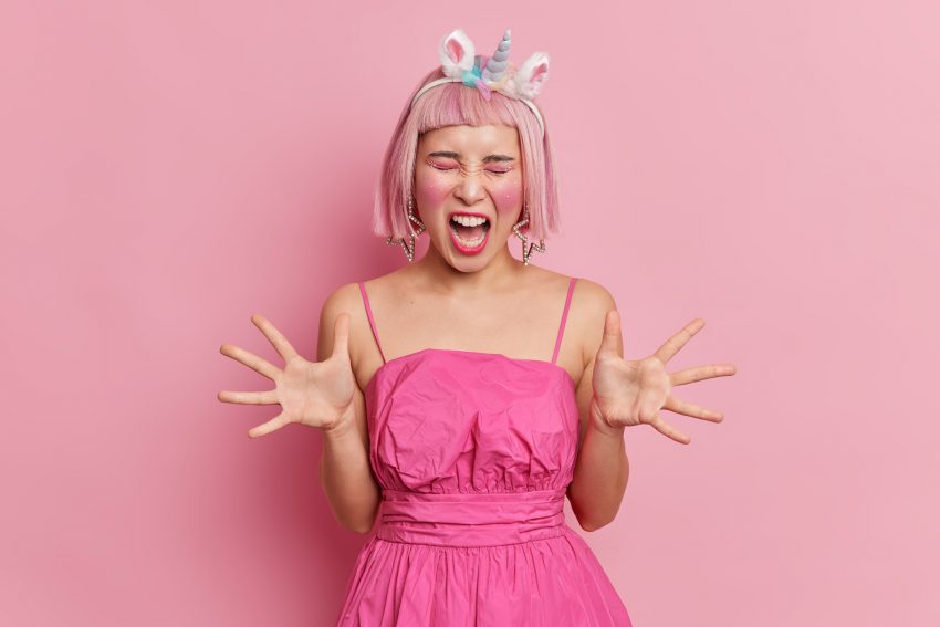 Emotional pink haired Asian woman with makeup and rhinestones on face raises palms screams angrily b